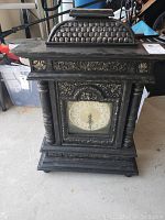 Front view of black ornate decorative tabletop clock with carved details and clock face visible.
