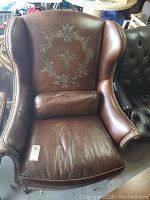 Front view of brown leather wingback chair with embossed backrest design and pillow on seat.