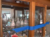Photo showing inside of wooden cabinet with multiple ceramic bells, glass and crystal figurines, and decorative items on glass shelves.