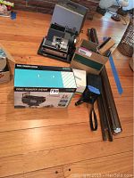 Vintage Bell & Howell projector with case, boxed video transfer system, DALITE screen poles and accessories arranged on hardwood floor.