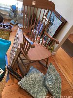 Wooden rocking chair shown with curved armrests, vertical back slats, light natural wood finish with a slightly contoured seat, placed near windows with other decor items behind.