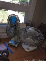 Three assorted vintage table top fans displayed near a window on wooden floor, showing varied designs and sizes, largest fan to left with blue blades and rectangular base.