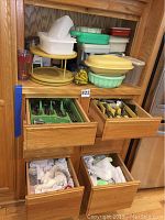 Plastic storage containers in various colors stacked on wood shelves with two drawers open below showing organized flatware and some cloth items in bottom drawers.