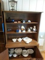 Wide shot showing wooden cabinet with shelves filled with items including clock on top shelf, glass art pieces, mugs, and china dishes in bottom cabinet.