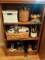 Full view of wooden shelf filled with office supplies such as baskets with pens, white plastic bin, stapler, and tape dispenser.