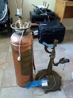 Photo shows two lamps side by side on a tiled floor. One lamp is tall and converted from copper/brass fire extinguisher with power cord and lamp fittings on top. The other lamp is made of heavy metal with ornamental design featuring multiple sockets for bulbs.