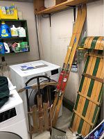 A pair of wooden vintage Shasta skis leaning against a wall near a washer and dryer, with a small wooden sled with metal runners next to them.