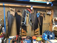 Image showing multiple hand saws hanging on pegboard including large wooden handled saws, metal saw frames with handles, and smaller saws. Various rust spots visible.