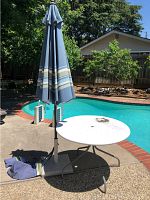 Photo of white outdoor round metal table with blue striped umbrella inserted, located beside pool