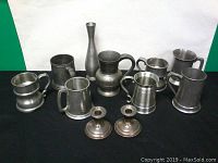 Photo showing nine assorted pewter items including tankards, mugs, a vase and two sterling silver candle holders displayed on black fabric.