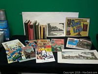 Assortment of books, comic books, sketches, and a colorful music sheet laid out on a black surface against a green background