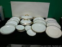 Full view of all plates and two lidded serving dishes with gold trim and handles displayed on a black surface.