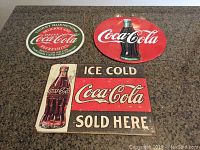 Three Coca-Cola replica metal signs arranged on a countertop including a circular green sign, a red oval sign with bottle, and a rectangular sign with bottle and text