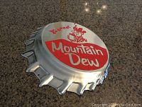 Photo showing a silver metal bottle cap shaped Mountain Dew replica sign with red and white logo and cartoon character.