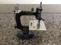 Front view of miniature Singer sewing machine showing black body, silver base, and Singer logo.