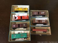 Seven HO scale model box cars in original boxes shown on a wood table. Cars include Canadian National, Great Northern, Schlitz, CP Rail, two ST maroon box cars, and a flatbed.
