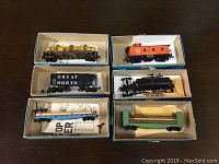 Six HO scale freight cars in their individual boxes seen from above, showing yellow Shell boxcar, orange caboose, black gondola, black tanker car, blue flatcar with wooden poles, and green flatcar with wooden load.