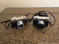 Front view of two Minolta Hi-Matic 11 cameras showing metal bodies, lenses, and one with lens cap.