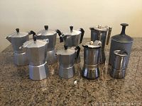 Front view of nine individual espresso pots arranged on a countertop, showing variety of sizes and one with broken handle.
