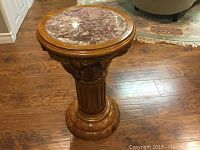 Full view of the round carved wood pedestal stand with marble top on a hardwood floor.