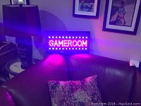LED sign showing the word GAMEROOM with red LEDs and blinking blue LED lights above and below the text, placed on a brown leather couch beside a table and lamp.