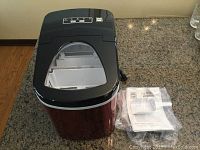 Front view of the portable RCA ice machine on a countertop with instruction card and scoop beside it in plastic wrapping.