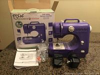 Purple Singer Pixie craft sewing machine with power adapters, threading tools, accessories, and instruction manual, original box in the background.