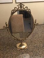 Front view of oval brass adjustable makeup mirror showcasing decorative filigree top and sides, mounted on a matching ornate stand, placed on granite surface with generic background.