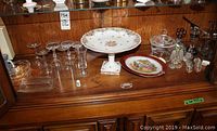 Multiple glassware and decor pieces arranged on wooden surface, including stemware, decorative plates, salt and pepper shakers, and clear glass dishes.