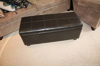 Dark brown faux leather rectangular storage bench with grid-stitched pattern on top and sides placed between beige couches on light carpeted floor.