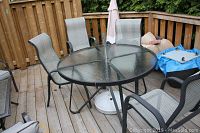 Wide shot of oval glass patio table with 4 metal-framed mesh chairs on a wooden deck, umbrella in center hole with no visible post, only fabric, and white umbrella base on floor