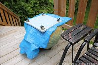 View of the large green ceramic planter covered partially by a blue tarp, placed on a wooden deck with a black metal dolly with wheels beneath it.