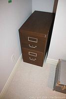Metal two-drawer filing cabinet with label holders and metal handles, showing scratches on top surface and sides, placed against a wall on carpeted floor.