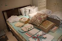 Photo showing assorted linens including folded napkins, decorative cushions with floral patterns, pillows, and a rolled comforter placed on a bed.
