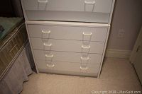 White laminate 4 drawer dresser shown against a wall near a bed, with visible plastic handles on each drawer.