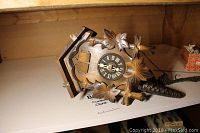 Wooden cuckoo clock with carved leaves and bird ornamentation, showing front clock face with Roman numerals and clock hands, and pine cone weight partially visible.