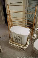Wooden clothes drying rack of light wood with wear. Plastic laundry hamper with lid and two plastic laundry baskets. Floor tile visible.