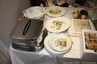 Multiple decor plates including floral patterned plates, Holly Hobbie plate, porcelain serving dish, and metal lidded cake plate on table with other small decor pieces and glassware.