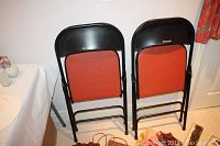 Two black Samsonite folding chairs with padded rust-orange backs, folded and leaning against a wall.
