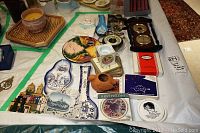 Photo showing assorted travel souvenir plates and coasters, decorative ceramic items, and barometer instruments on a table covered with white cloth.