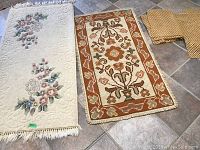 Two small carpets on tile floor. Larger cream colored floral oriental hand-knotted rug with fringes and smaller orange and brown geometric floral carpet with looped edges.