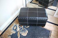 Front view of the black padded storage bench with white stitching and metal studs on top edge, sitting on floral rug near mirrored closet door.