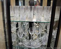 Photo showing 8 tall crystal tumblers on the upper shelf and assorted glasses and serving pieces on the lower shelf in a display cabinet.