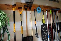 Wide view of garden tools hanging on wooden rail showing shovels, rakes, claw weeder, and green power cord.