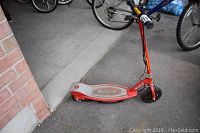Red Razor E100 motorized scooter leaning against a brick wall, showing the left side and front wheel.