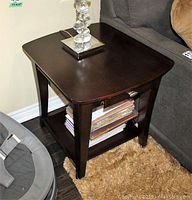 Pair of dark wood side tables shown together by a couch, each with a drawer and bottom shelf containing stacked papers and books. Minor surface wear visible.