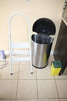 Trash can with open black lid next to white metal step stool on tile floor near wall outlet and electric plug.