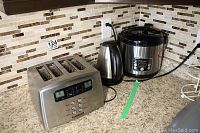 Photo showing three small appliances on a kitchen counter, a stainless steel 4-slice Cuisinart toaster, a black and stainless steel rice cooker, and a stainless steel electric kettle wired in.