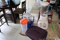 Photo showing Oster blender, Hamilton Beach mixer with transparent container attachment, orange shaker bottle, and purple Tupperware container on kitchen counter.