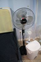 Black Garrison pedestal fan next to bed and white trash can, showing condition and size (lstimg_01j8xbdhrhfh7tj19sek2ah7jv)